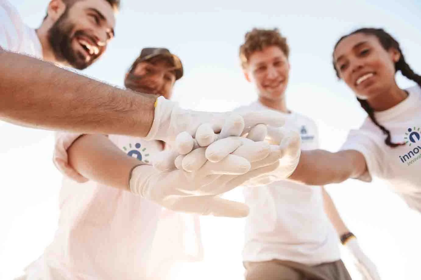 Four young volunteers in white T-shirts and white work gloves stacking their hands together in a circle, seen from below against a bright sky